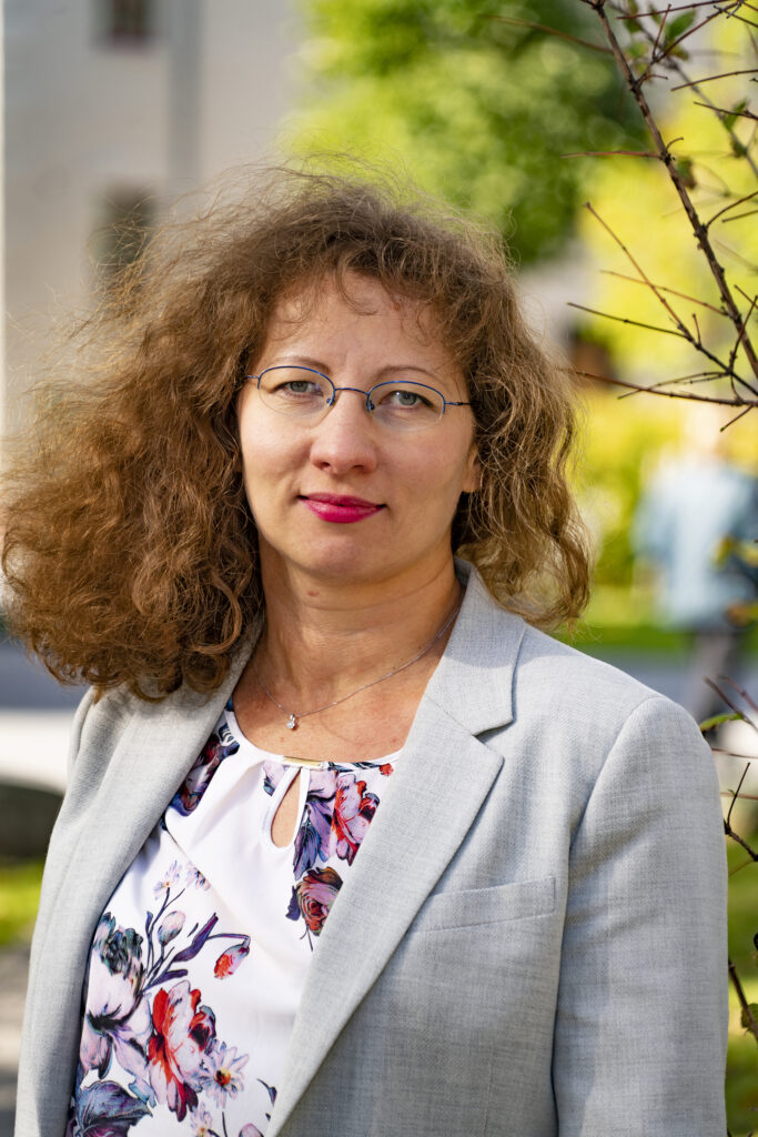 Portrait of a woman with curly hair wearing glasses and a light grey blazer, standing outdoors.